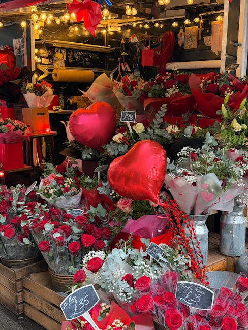 An indoor floral display featuring an assortment of fresh red and white flowers, including roses and carnations, arranged in baskets and wrapped bouquets. Prominent are two large, shiny red heart-shap