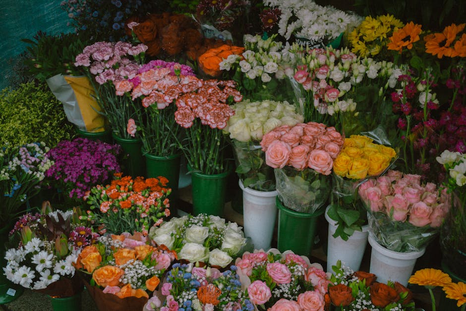 A vibrant display of fresh flower bouquets and potted plants arranged on shelves and the floor of a flower shop, featuring a variety of flowering plants and cut flowers including pink, white, yellow,