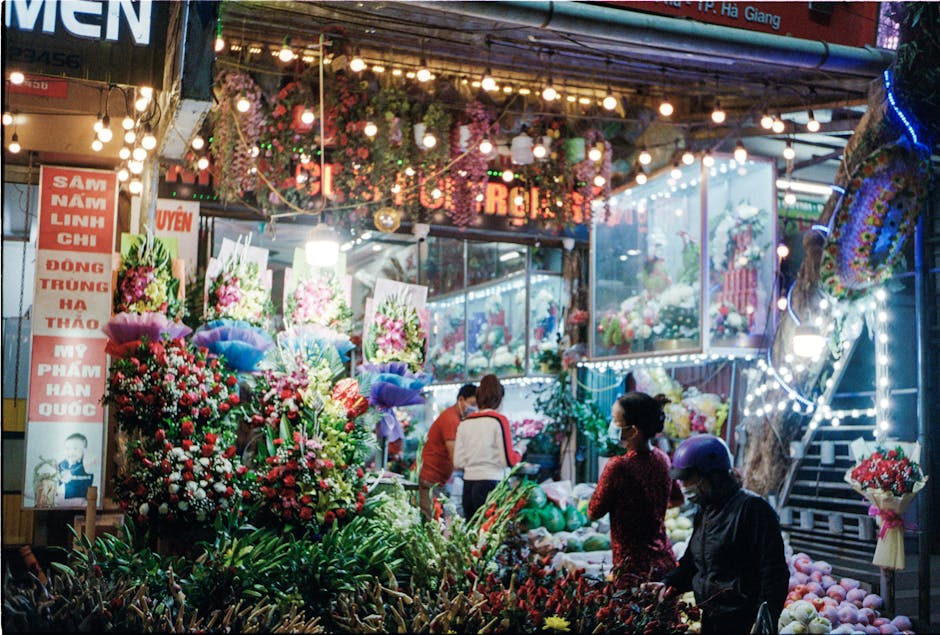 A vibrant flower stall at night illuminated by string lights, featuring an array of fresh bouquets and floral arrangements in various colors, predominantly reds, pinks, and whites. The display include