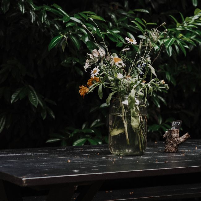 A glass jar filled with a casual bouquet of fresh wildflowers, including white daisies with yellow centers, orange blooms, and delicate green foliage, placed on a dark wooden table. The flowers appear